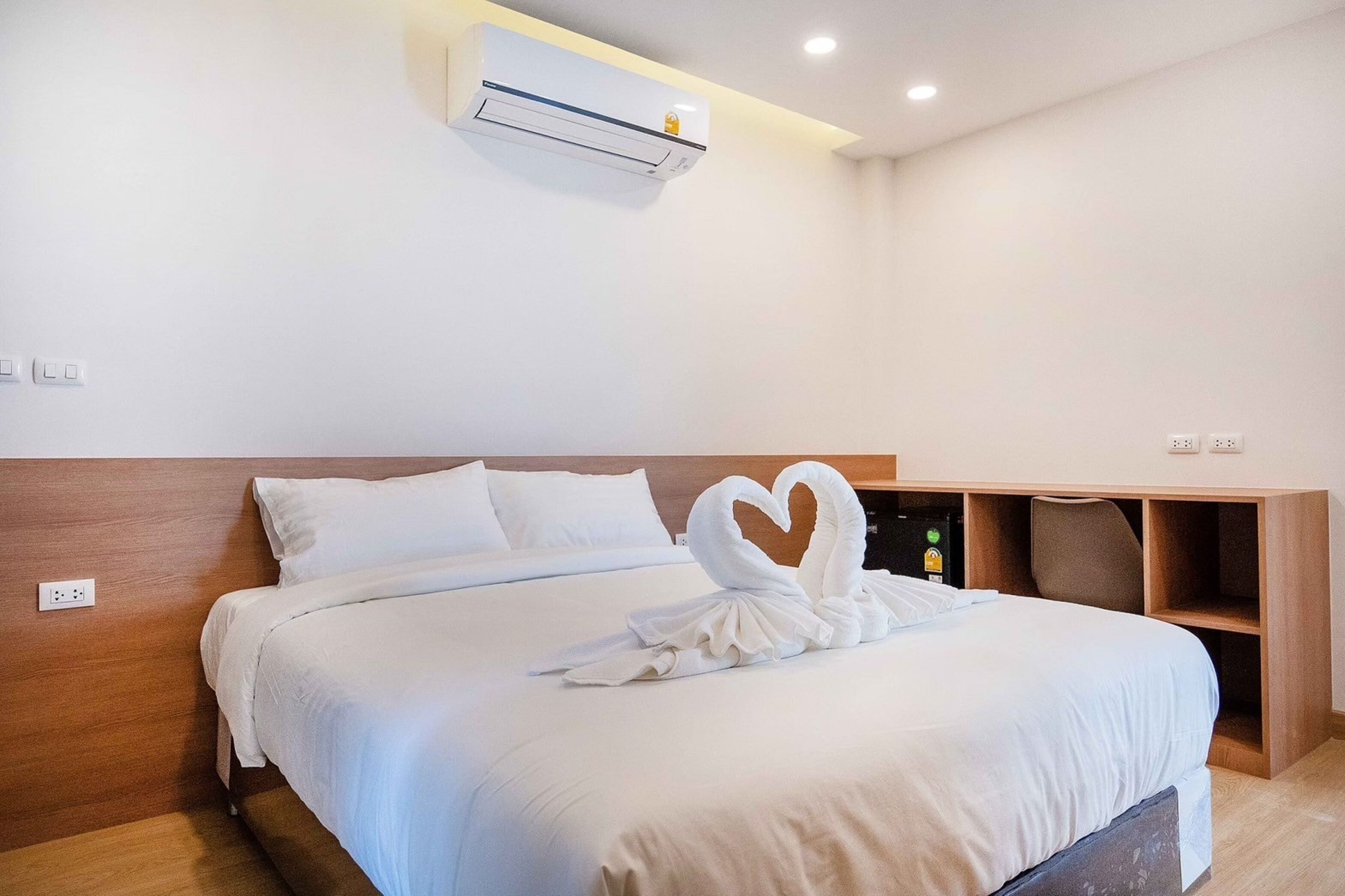 A hotel room with a neatly made bed, a heart-shaped towel on the bed, a built-in shelf, and a wall-mounted air conditioner.