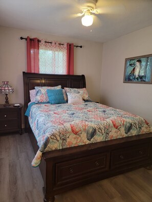 A bedroom with a wooden bed, a nightstand with a lamp, a window with patterned curtains, and a painting on the wall.