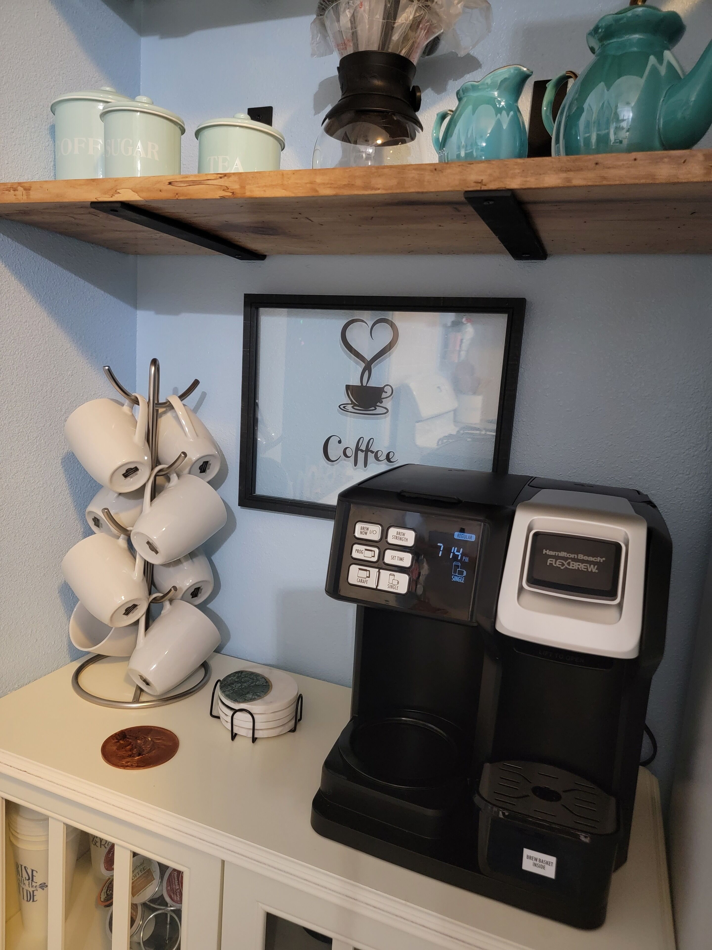 A coffee machine with a digital display, a coffee mug rack, and a framed coffee-themed artwork on a wall.