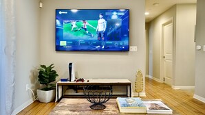 Un saló amb televisió plana que mostra un partit esportiu, una taula de centre de fusta amb llibres i un objecte decoratiu, una planta en test i una consola de jocs.