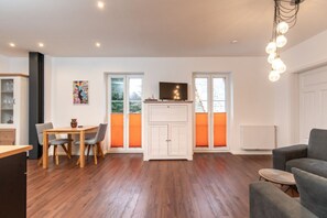 Een moderne woonkamer met een eethoek, houten vloeren, een televisie op een kast en grote ramen voorzien van oranje luiken.