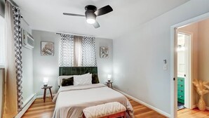 Ein Schlafzimmer mit Bett, Deckenventilator, Fenster mit Vorhängen, Nachttisch und Beistelltisch.