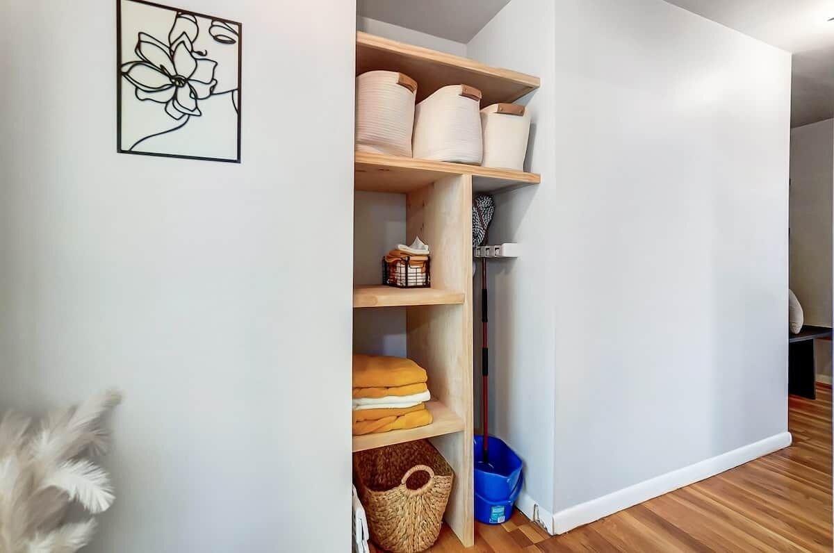 A hallway with a white wall, a framed artwork, wooden shelves with storage bins, and a blue trash bin.