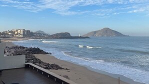 Vista sulla spiaggia con un litorale roccioso, un faro in lontananza e un edificio con un balcone affacciato sull'oceano.