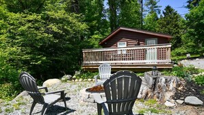A cabin with a deck, surrounded by trees and a fire pit area with chairs. - Close to Town - Pet Friendly Cabin w/ Fire Pit (Lake George)