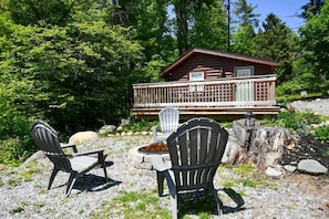 A cabin with a deck, surrounded by trees and a fire pit area with chairs. - Close to Town - Pet Friendly Cabin w/ Fire Pit (Lake George)
