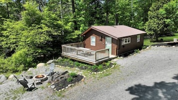 Une cabane dotée d’une terrasse, entourée d’arbres, et un foyer avec des chaises.