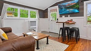 A living room with a brown leather sofa, a wooden coffee table, and a TV mounted on the wall. - Close to Town - Pet Friendly Cabin w/ Fire Pit (Lake George)