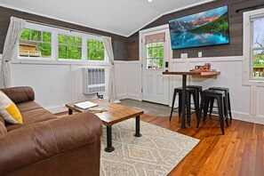 A living room with a brown leather sofa, a wooden coffee table, and a TV mounted on the wall. - Close to Town - Pet Friendly Cabin w/ Fire Pit (Lake George)