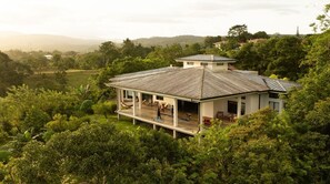 A house with a large porch, surrounded by lush greenery and hills in the background.
