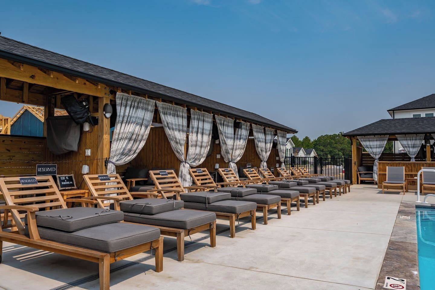 A row of wooden lounge chairs with cushions, a poolside area, and a building with a wooden facade and curtains.