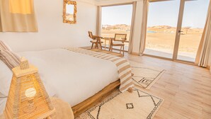 Una habitació àmplia amb un llit gran, taula de menjador amb caires i vista al desert a través de grans finestres.