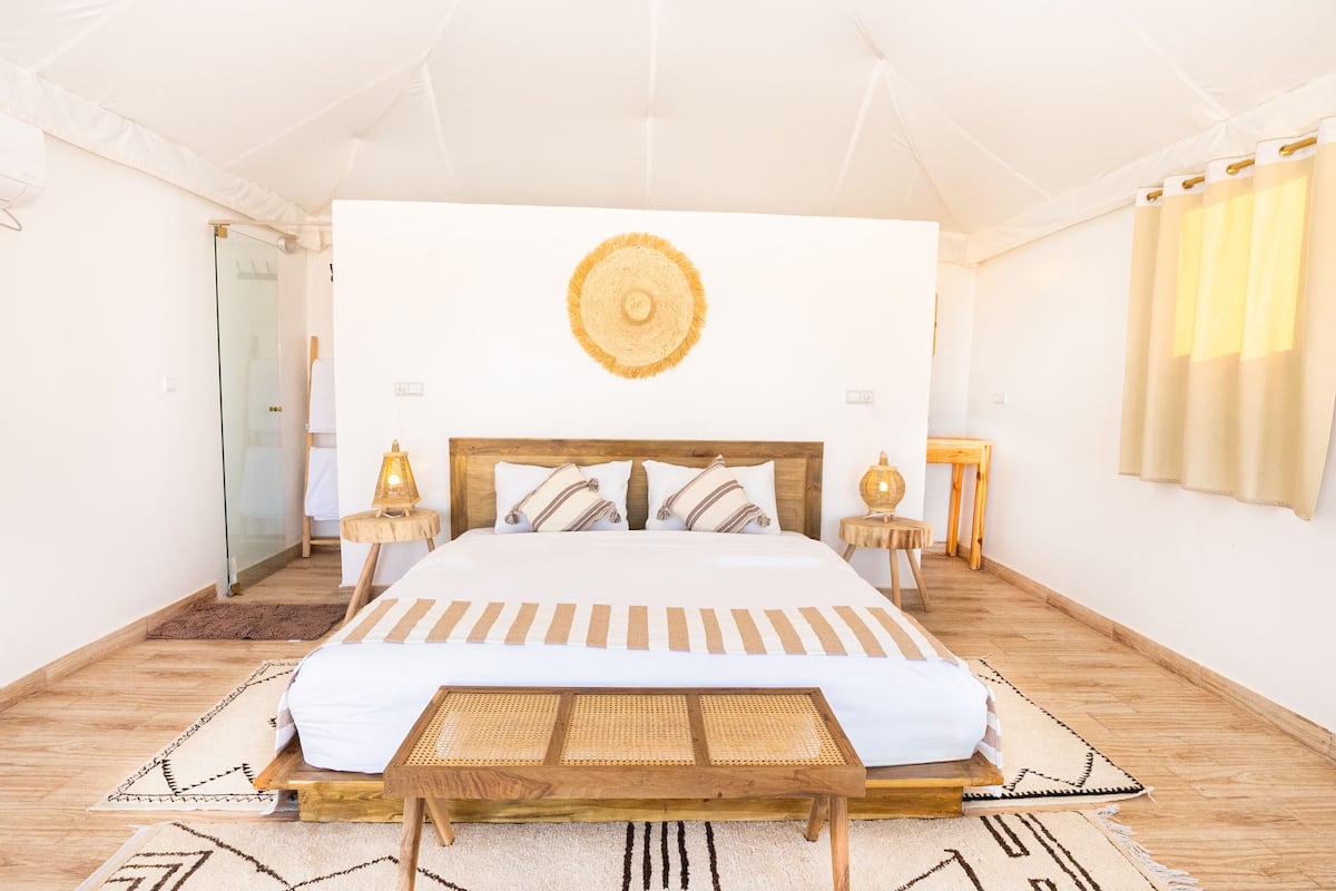 A spacious bedroom with a large bed, a wooden headboard, a wall-mounted circular light fixture, and a glass door leading to a bathroom.