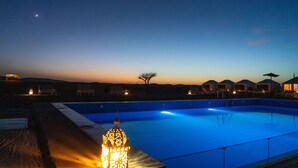 Una piscina al capvespre amb un cel clar, una lluna en forma de creixent i alguns edificis al fons.