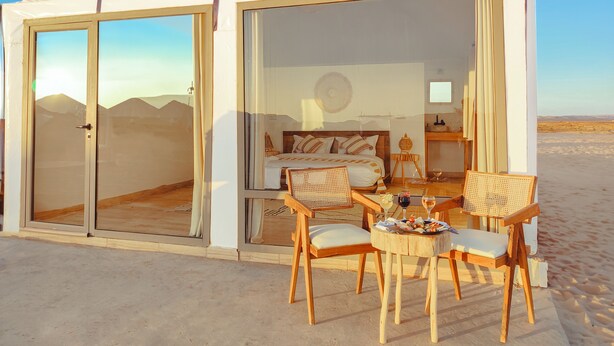 Un allotjament en tenda amb un llit, dues cadires, una taula amb una copa de vi i vista al desert.