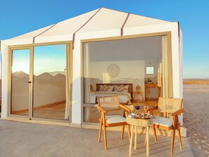 A tented accommodation with a bed, two chairs, a table with a wine glass, and a view of the desert. - Sahara Royal Resort (Rissani)