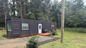 Ein kleines, eingeschossiges Blockhaus mit einer kleinen Veranda, umgeben von einem angelegten Garten und einem bewaldeten Bereich.