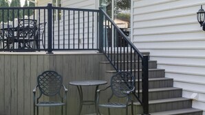 A patio with two metal chairs and a small round table, a wooden deck, and a set of stairs leading to a white building.
