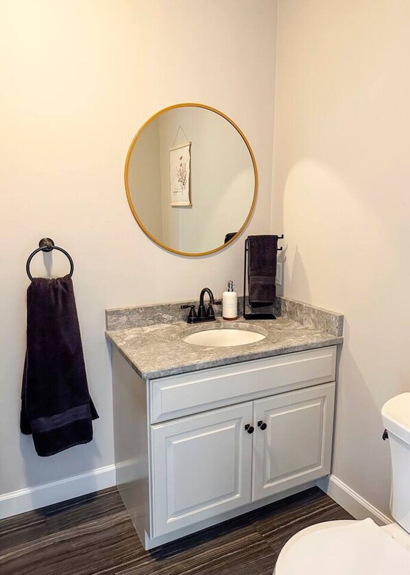 Une salle de bain avec un lavabo blanc, un miroir rond doté d’un cadre doré, une serviette noire et des toilettes blanches.