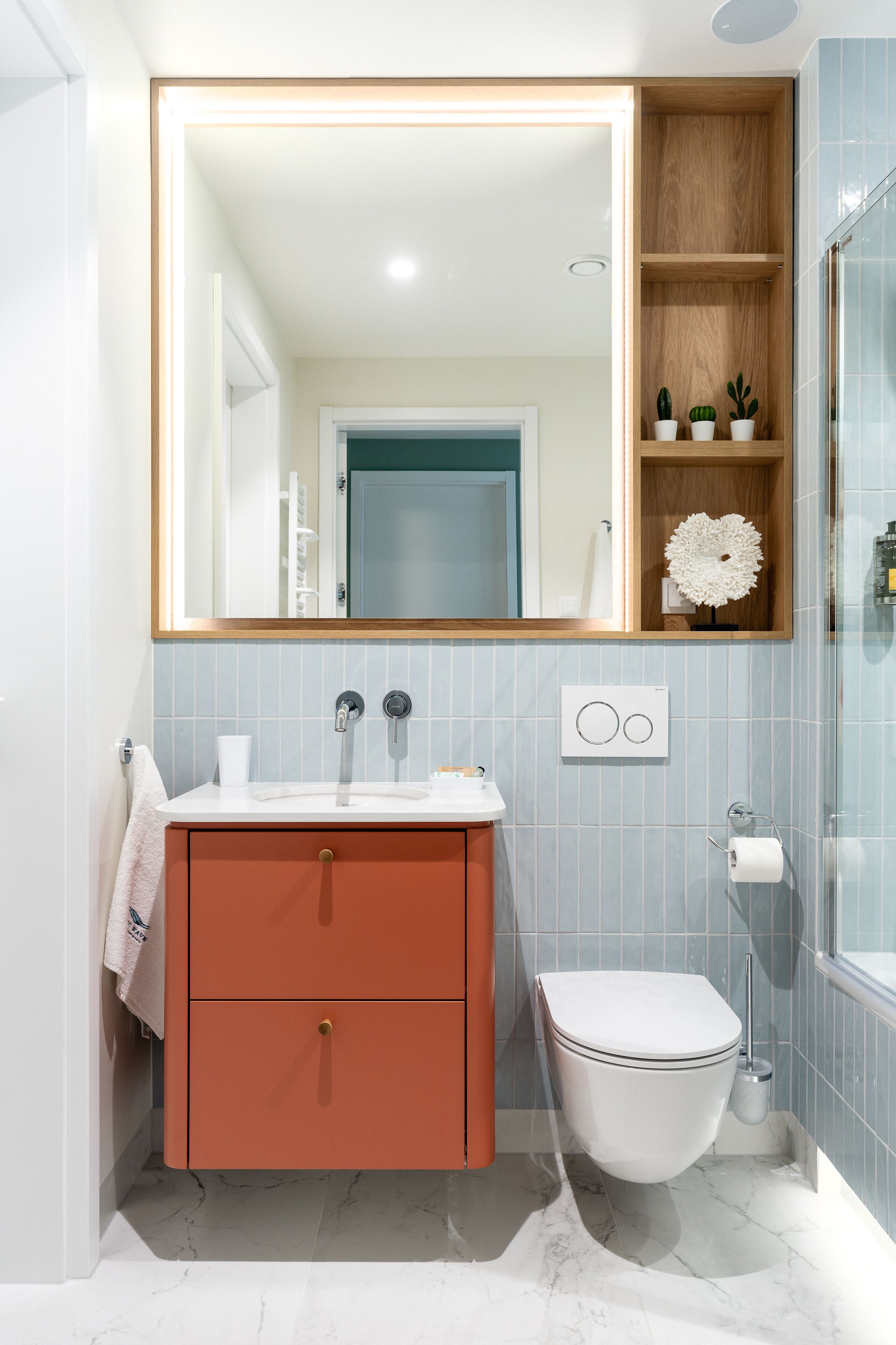 Un baño con inodoro blanco, un lavamanos con espejo y una repisa de madera con plantas.