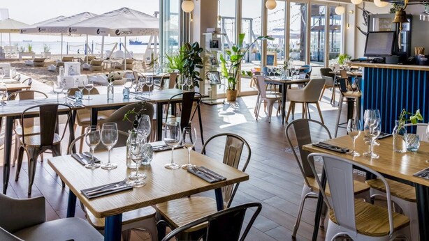 Restaurante com área externa e mesas e cadeiras de madeira, oferecendo vista para a praia e grandes janelas com vista para o mar.