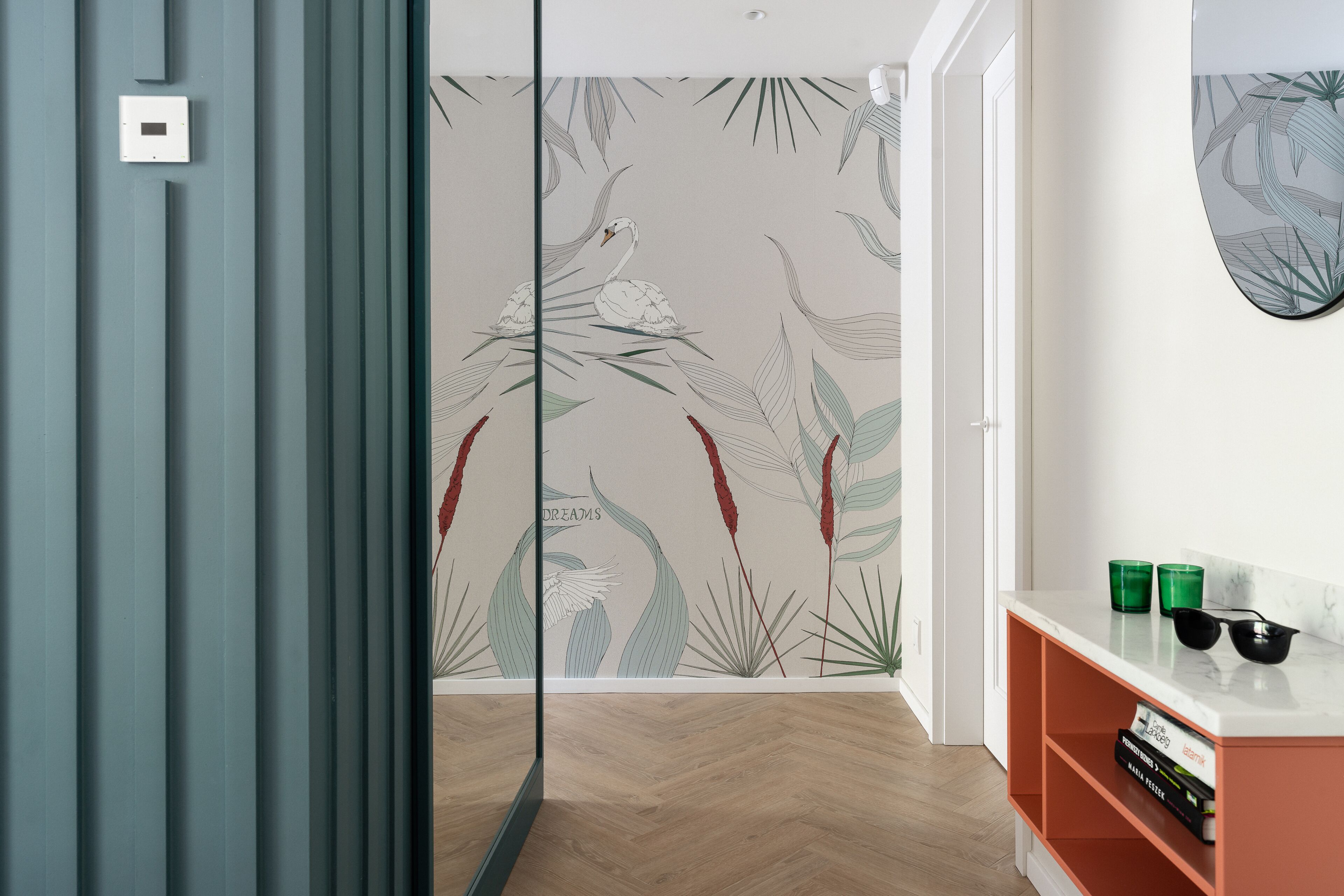 Una habitación moderna con suelo de madera tipo espiga, pared blanca con estampado floral, separador de vidrio y estante blanco con dos tazas verdes y un par de gafas de sol.