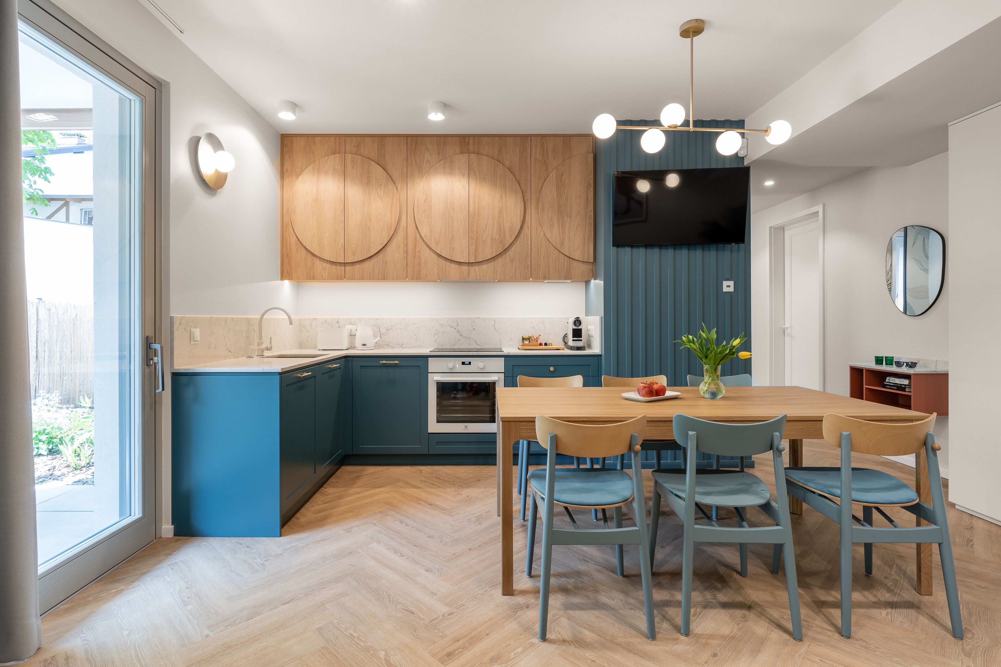 A modern kitchen with blue cabinets, a wooden dining table, and a TV mounted on the wall.