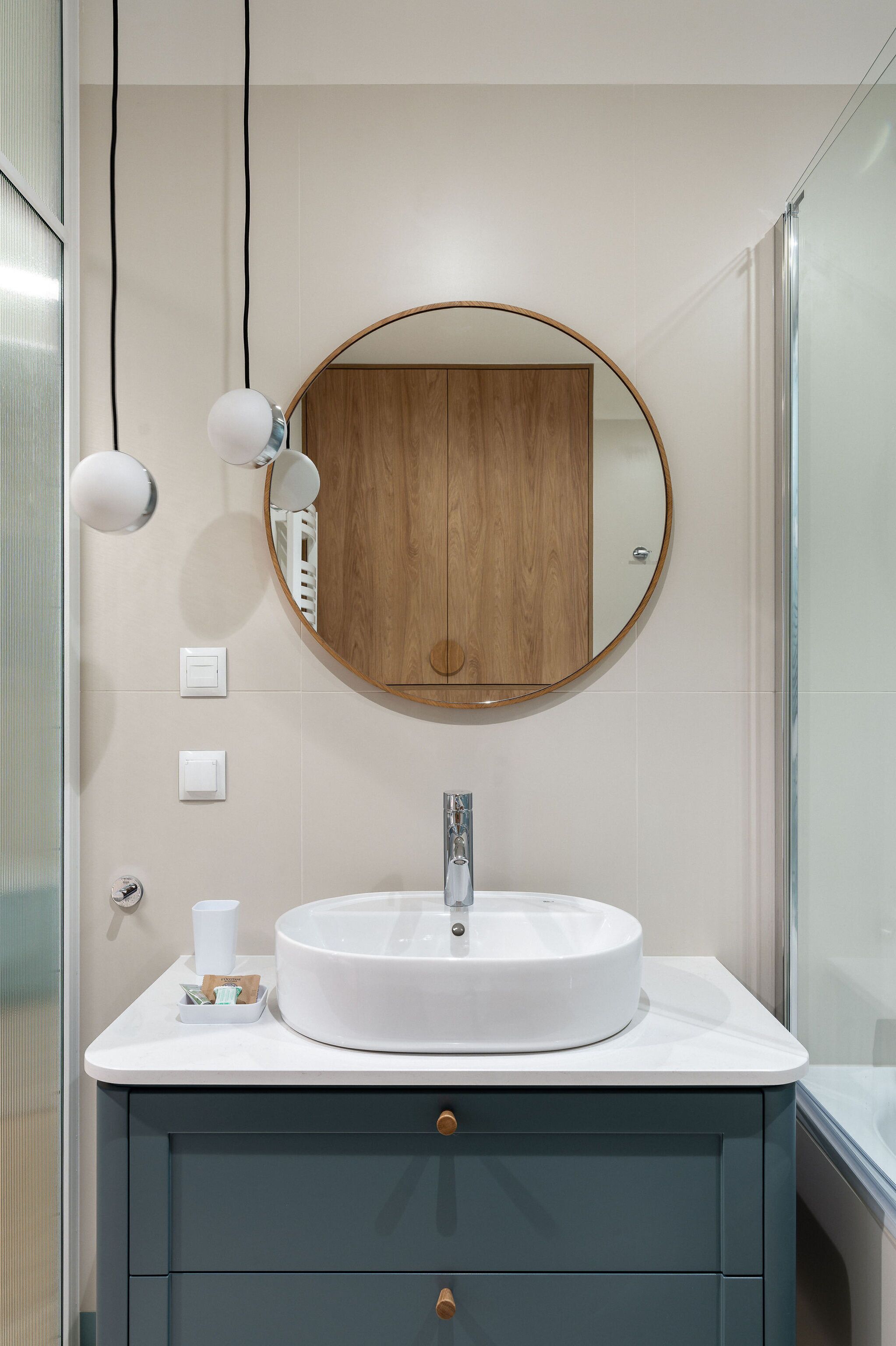 Un baño con un espejo redondo, lavamanos blanco y un mueble de madera.