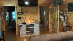 A wooden cabin interior with a TV, a small table with a lamp, a bookshelf, and a couch.