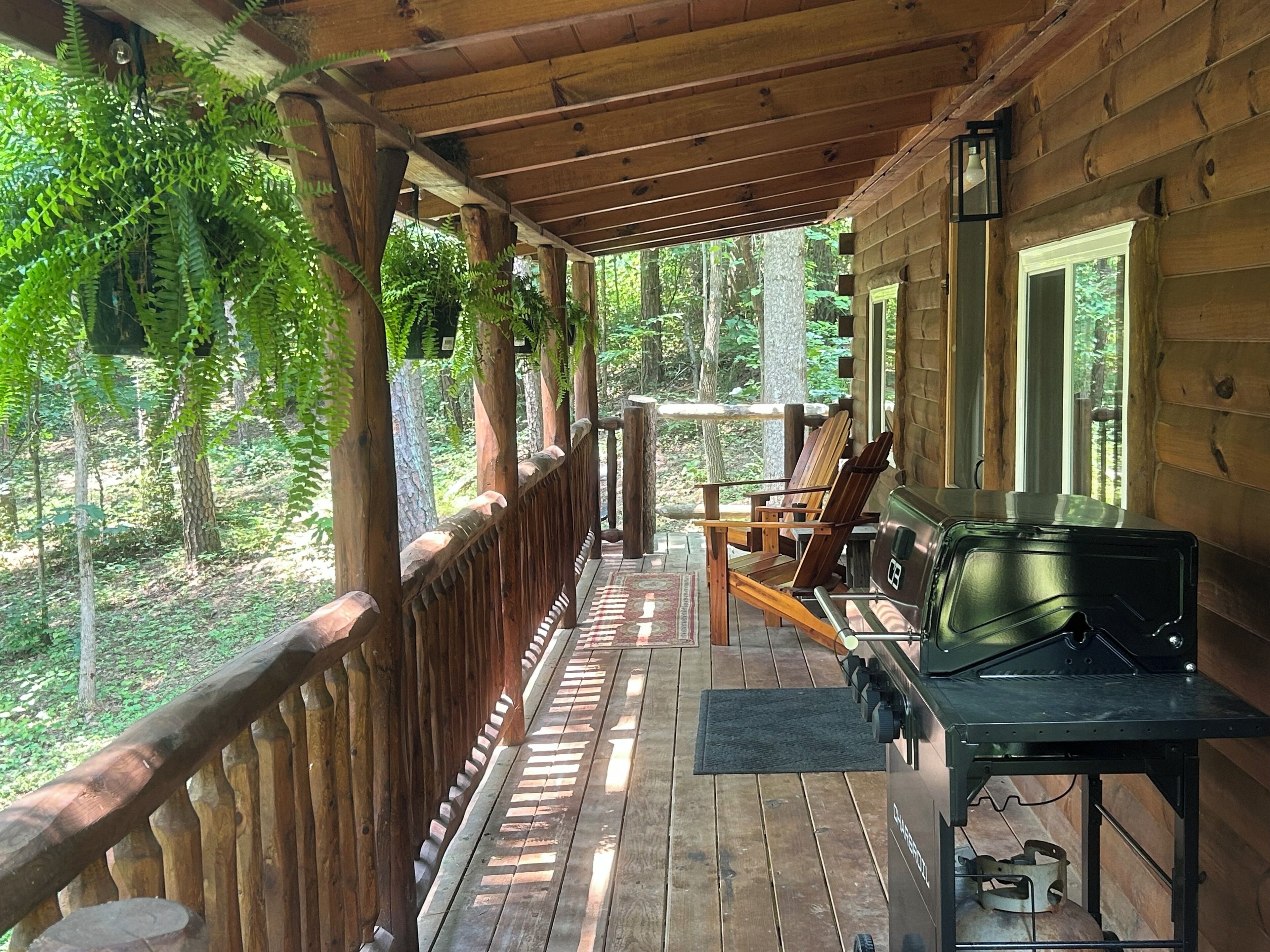 A wooden porch with a grill and chairs, surrounded by trees.