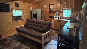 A cozy cabin interior with a sofa, kitchenette, wooden walls, and a ceiling with exposed beams.