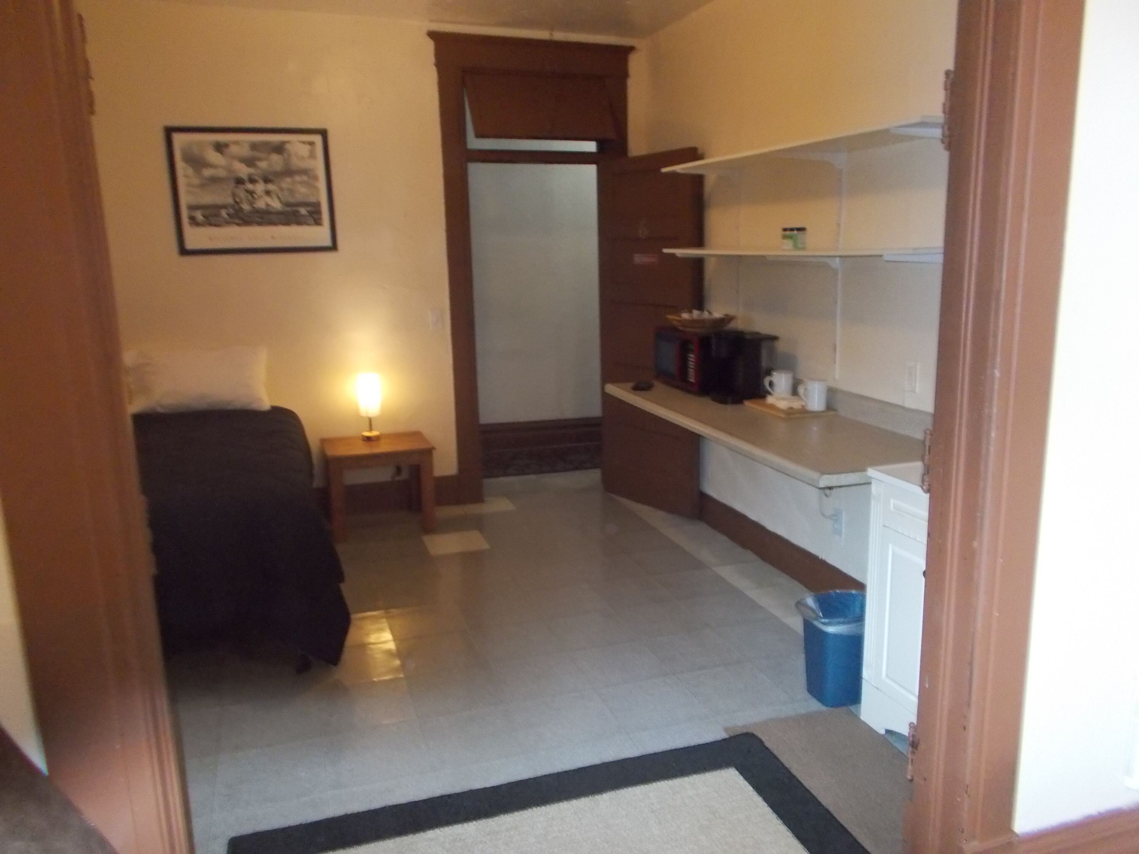 A compact hotel room with a bed, a bedside table with a lamp, a kitchenette with a microwave and coffee maker, and a framed picture on the wall.