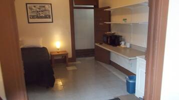 A compact hotel room with a bed, a bedside table with a lamp, a kitchenette with a microwave and coffee maker, and a framed picture on the wall.