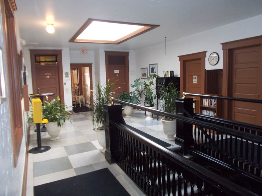 A hotel lobby with a checkered floor, a staircase with a black railing, and several potted plants.