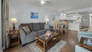 Un salon avec un canapé, une table basse et des fauteuils, un coin repas avec une table et des chaises, et une cuisine à l’arrière-plan.