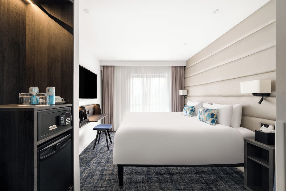 A hotel room with a large bed, a desk with a chair, a TV, and a minibar.
