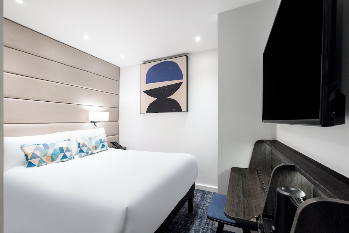 A hotel room with a bed, a desk, a chair, a TV, and a wall-mounted artwork.
