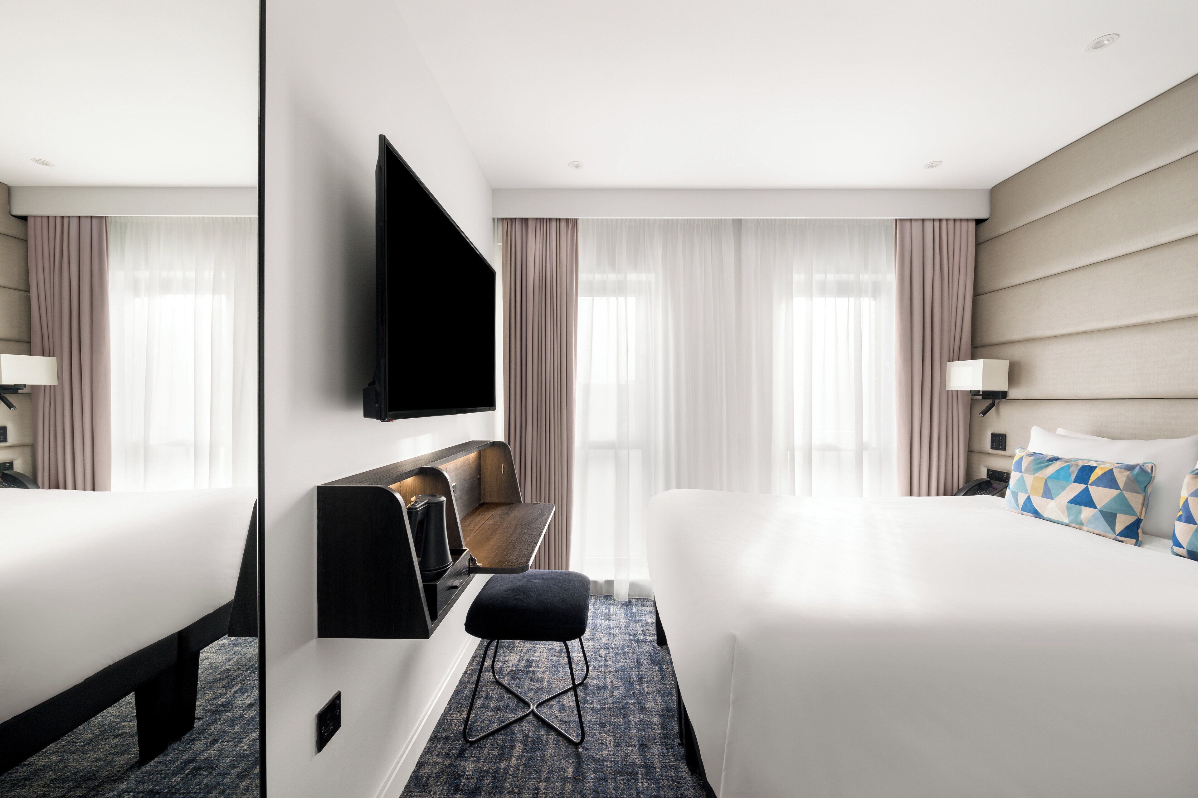 Una habitació d'hotel moderna amb un llit gran, una televisió de pantalla plana penjada a la paret, un escriptori petit amb una cadira i una finestra amb cortines transparents.