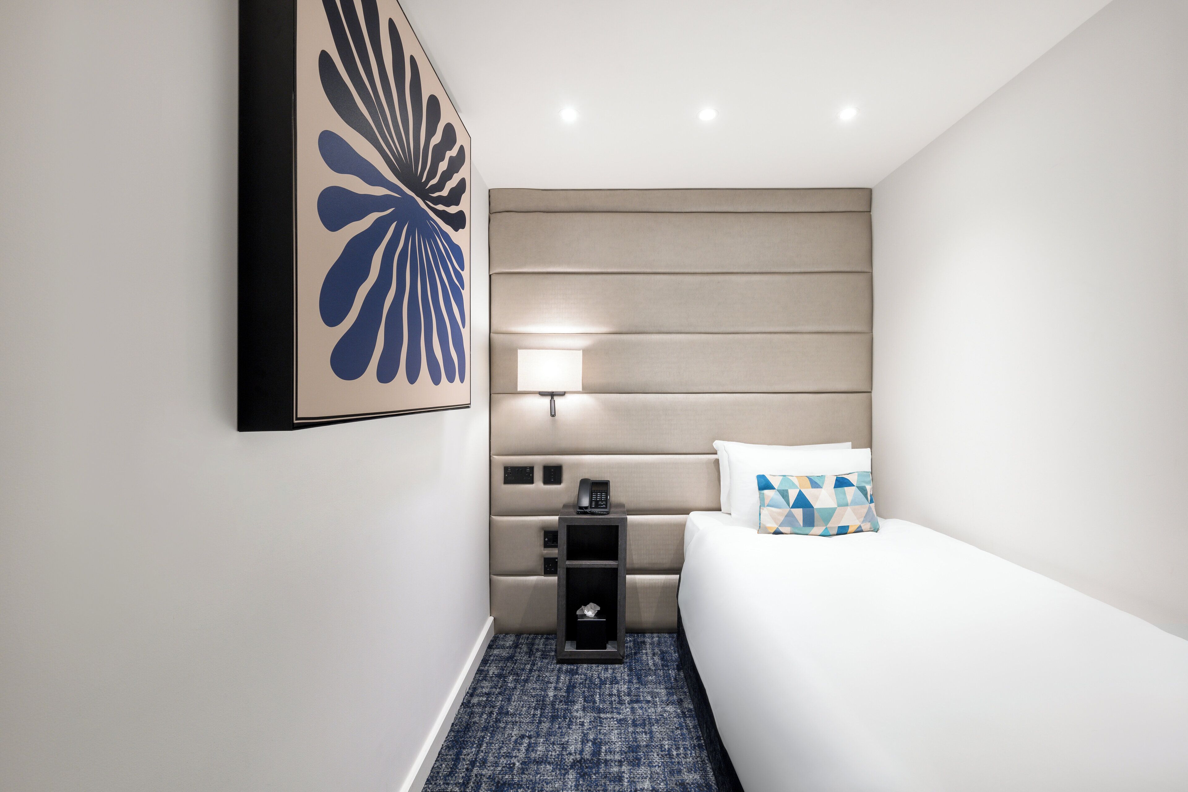 A hotel room with a bed, a bedside table with a lamp, a phone, and a decorative wall piece.