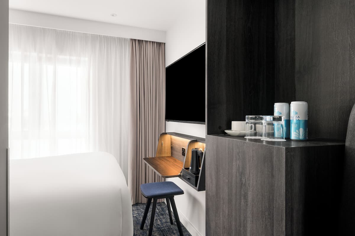 A hotel room with a bed, a desk with a chair, a coffee maker, and a shelf with cups and a water bottle.