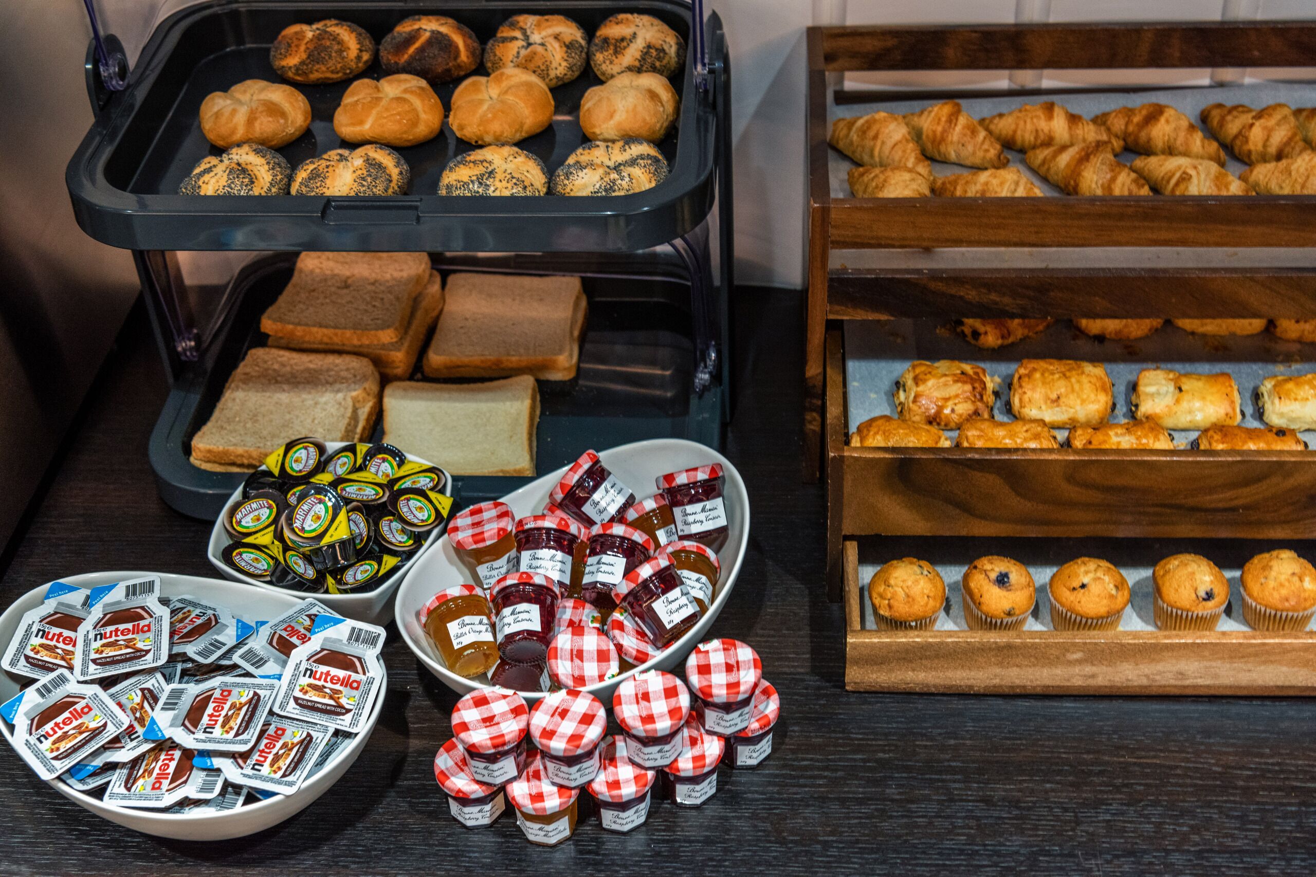 A buffet with various baked goods, including bread, pastries, muffins, and Nutella packets.