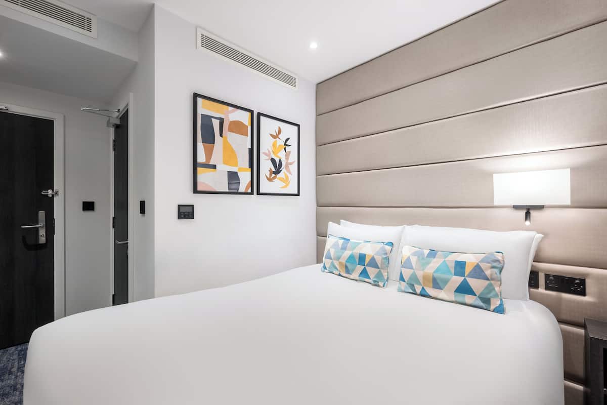 A hotel room with a large bed, two pillows, a headboard with controls, and two framed artworks on the wall.