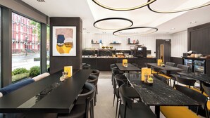 A modern restaurant with black and yellow tables, a kitchen in the background, and a view of a red building outside. - voco London Marylebone by IHG (London)