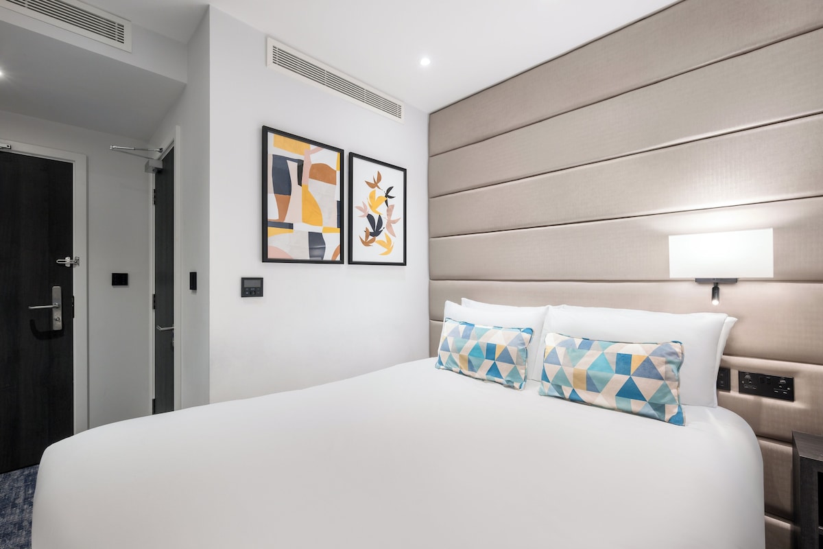 A hotel room with a large bed, two pillows, a headboard with controls, and two framed artworks on the wall.