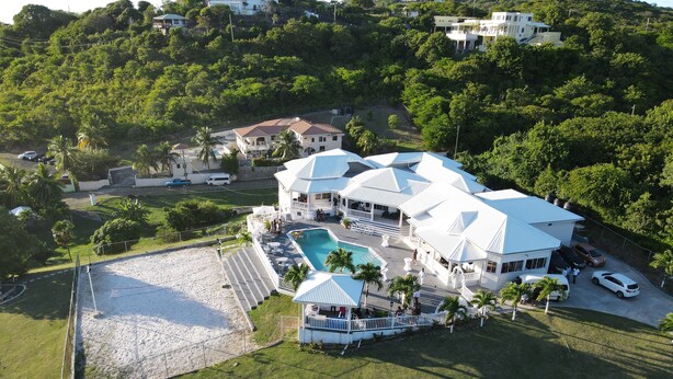 An aerial view of a large, white, multi-story house with a swimming pool, surrounded by lush greenery and a sandy area.