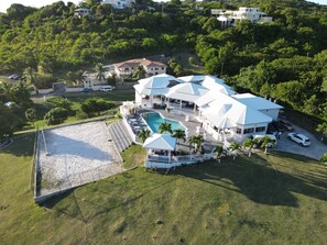 An aerial view of a large, white, multi-story house with a swimming pool, surrounded by lush greenery and a sandy area. - Paris Villas Cap Estate (Gros Islet)