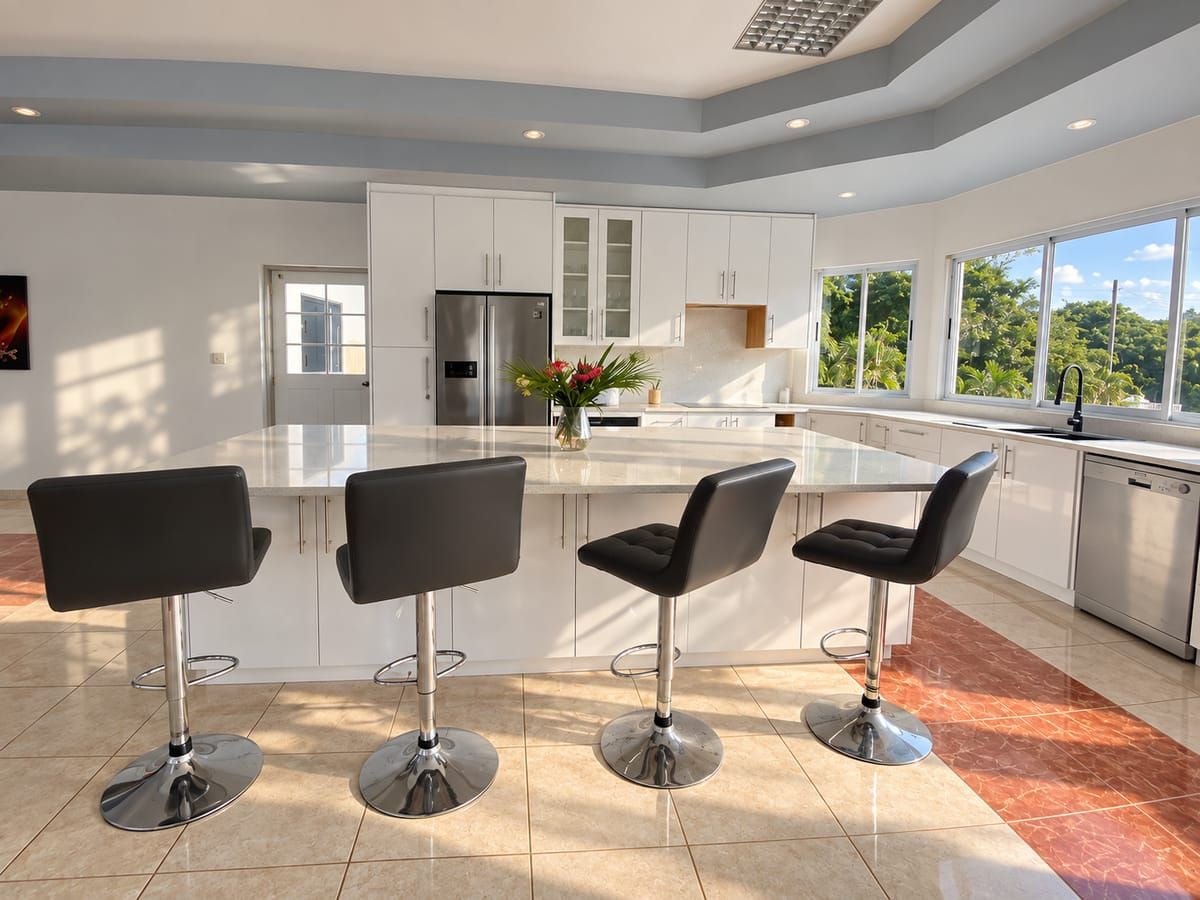 A modern kitchen with a central island, three bar-style chairs, and a view of greenery outside.