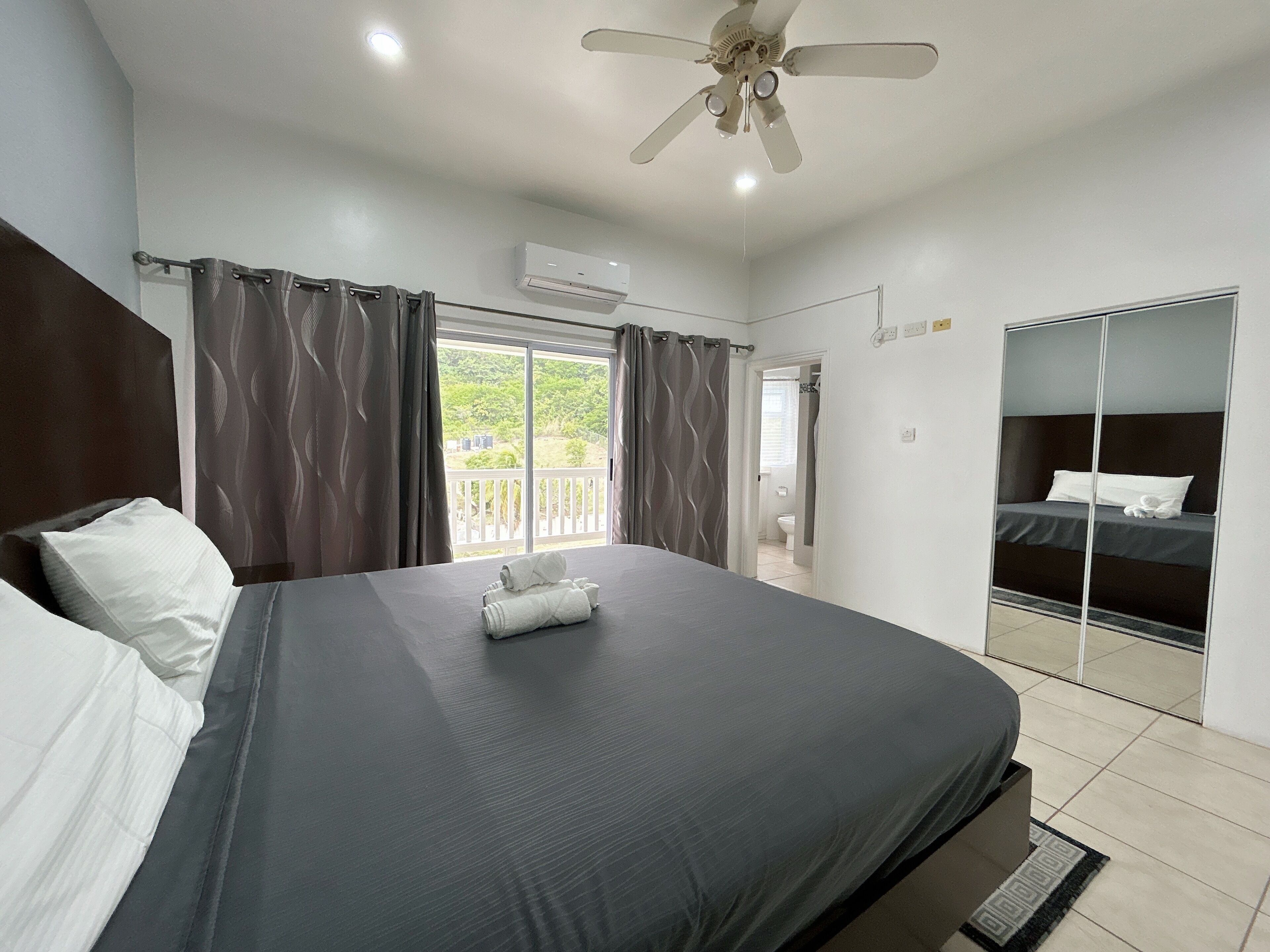 A bedroom with a bed, a ceiling fan, an air conditioning unit, a bathroom visible through an open door, and a balcony with a view of greenery.
