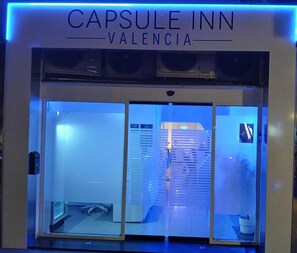 Exterior del Capsule Inn Valencia, con un diseño moderno, que presenta una entrada de vidrio y un letrero de neón azul.