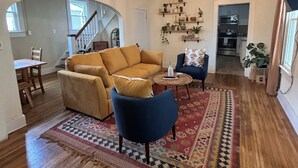 Un vaste salon comprenant un grand canapé, un fauteuil bleu et un tapis à motifs. On y trouve un escalier menant à un autre niveau, un coin repas et, au fond, une cuisine.